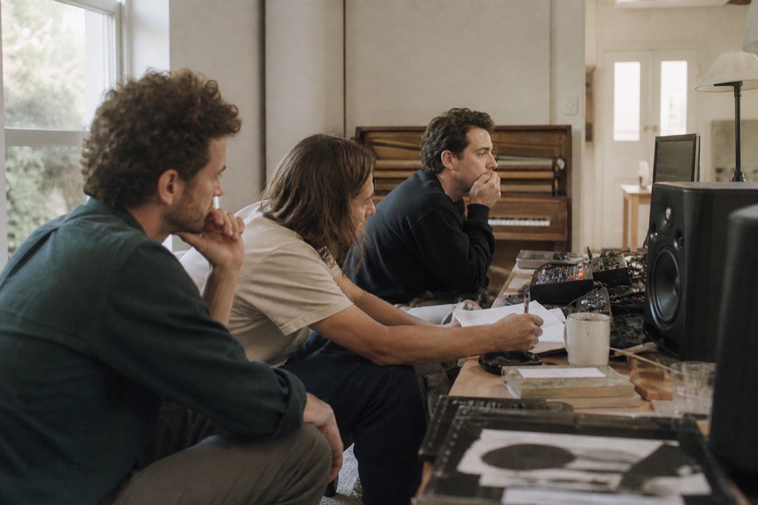 Three musicians listening at studio monitors in a daylit room, one taking notes