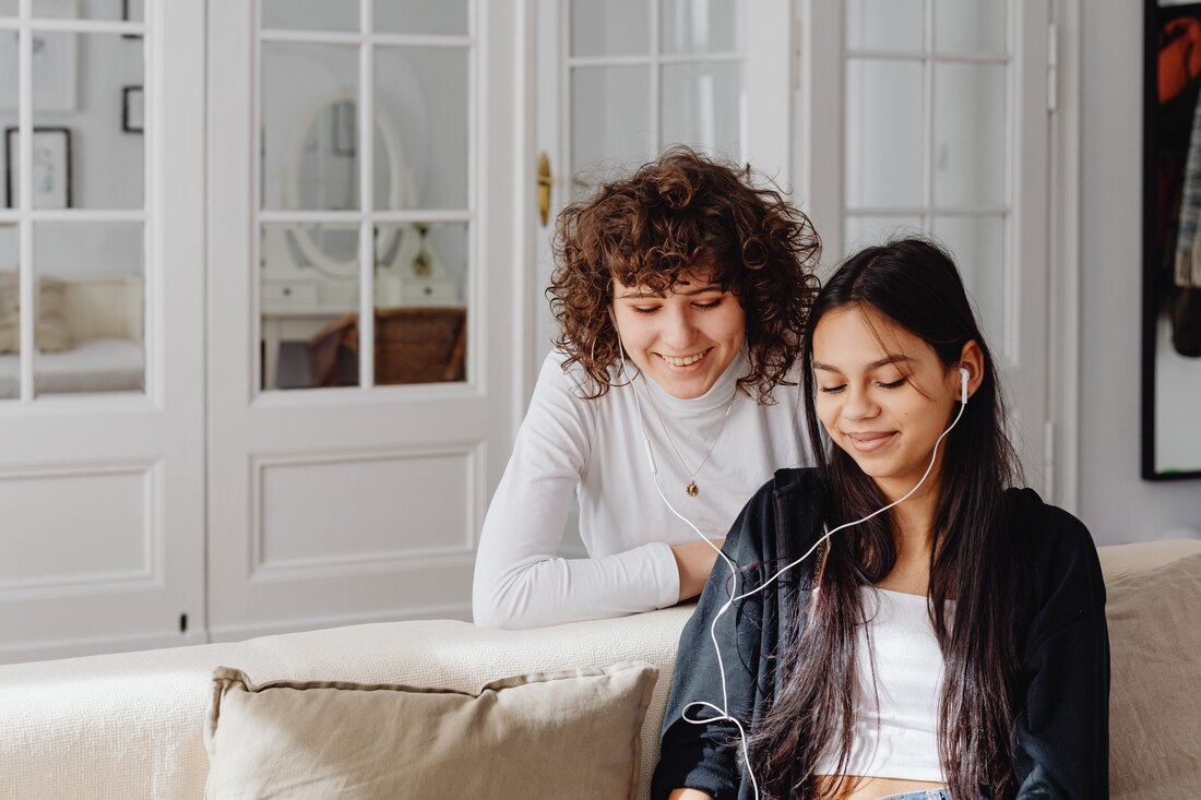 Two people on a sofa in a daylit room sharing a pair of earphones