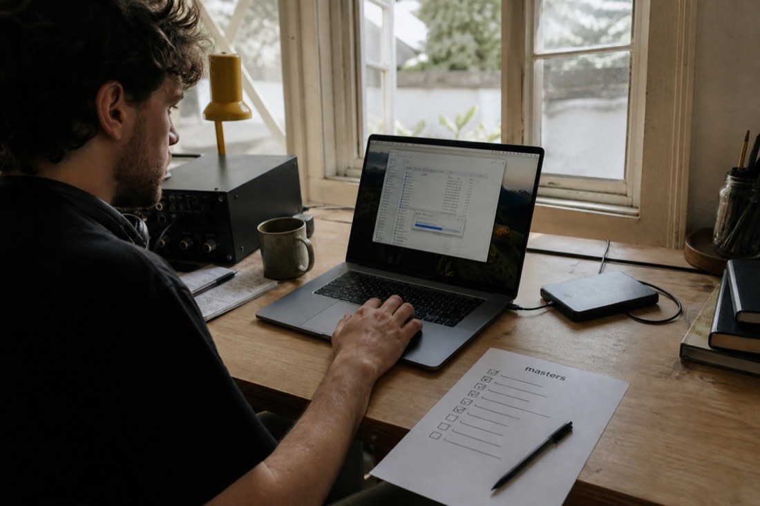 A musician at a desk in afternoon light, moving files from a laptop to an external drive, with a printed checklist titled masters, half checked off