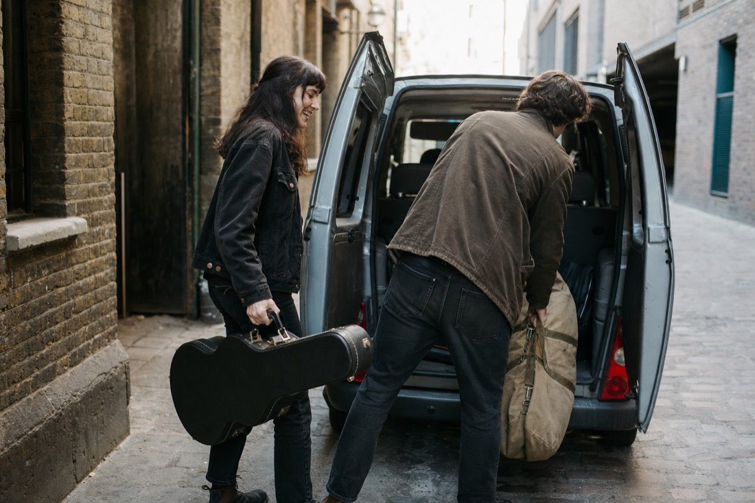 Two musicians carrying gear from a van through a daylit doorway